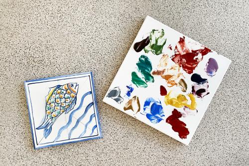 Piastrella di ceramica dipinta a mano che rappresenta un pesce, accanto piastrella con i colori usati durante il corso di pittura su ceramica in Lombardia