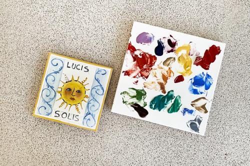 Una piastrella di ceramica dipinta a mano che raffigura un sole e delle decorazioni, vicino una piastrella con i colori utilizzati