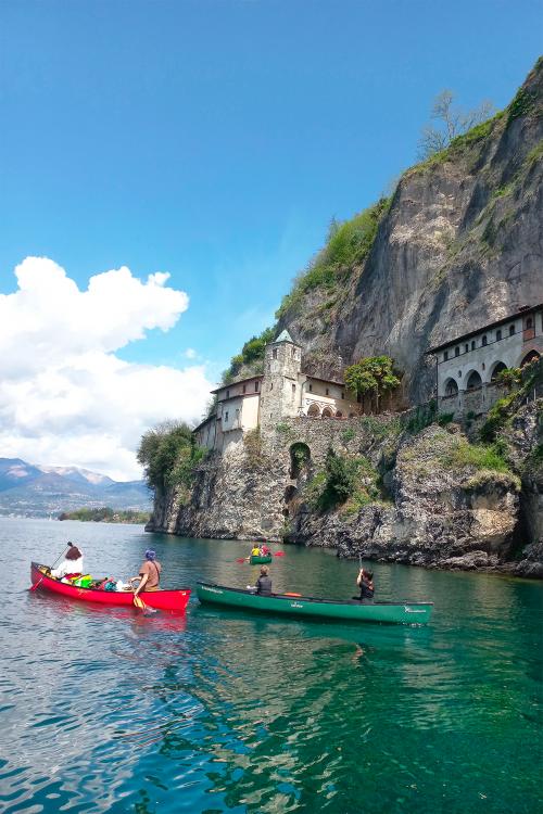 Delle persone a bordo di canoe canadesi pagaiano sul Lago Maggiore verso l'Eremo di Santa Caterina del Sasso
