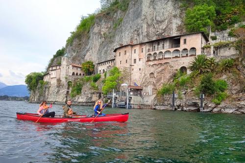 Tre ragazzi a bordo di una canoa canadese pagaiano sorridenti verso l'Eremo di Santa Caterina del Sasso sul Lago Maggiore